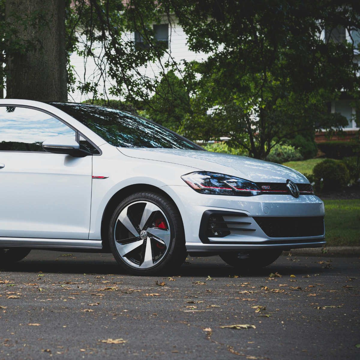 A white Volkswagen Golf GTI parked on a residential street, shot from a low front-quarter angle with lush green trees and a house visible in the background.