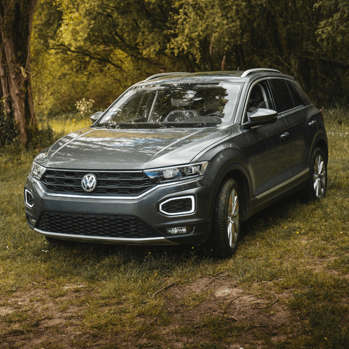 A grey Volkswagen T-Roc SUV parked on a grassy woodland clearing, shot from a straight-on front angle surrounded by dense green trees.