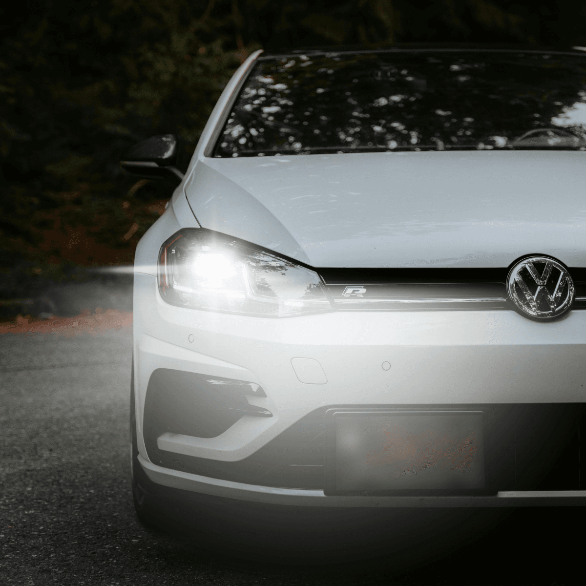 A close-up front view of a white Volkswagen Golf R with its headlights on, shot at night against a dark background of trees.