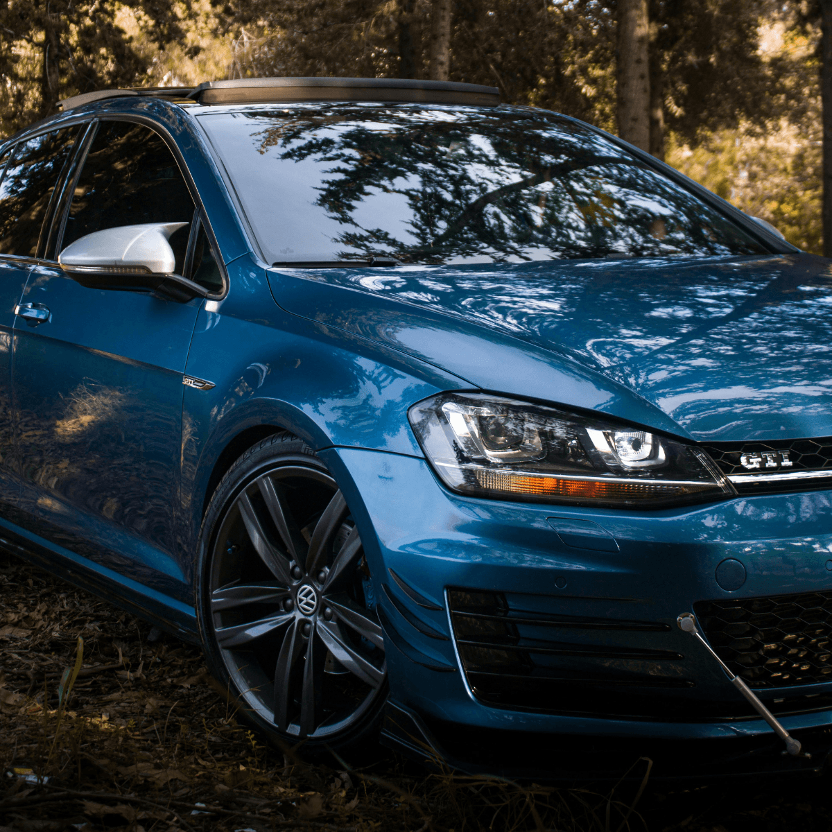 A blue Volkswagen Golf GTI parked among trees, shot from a low front-quarter angle with dappled sunlight highlighting the bodywork.