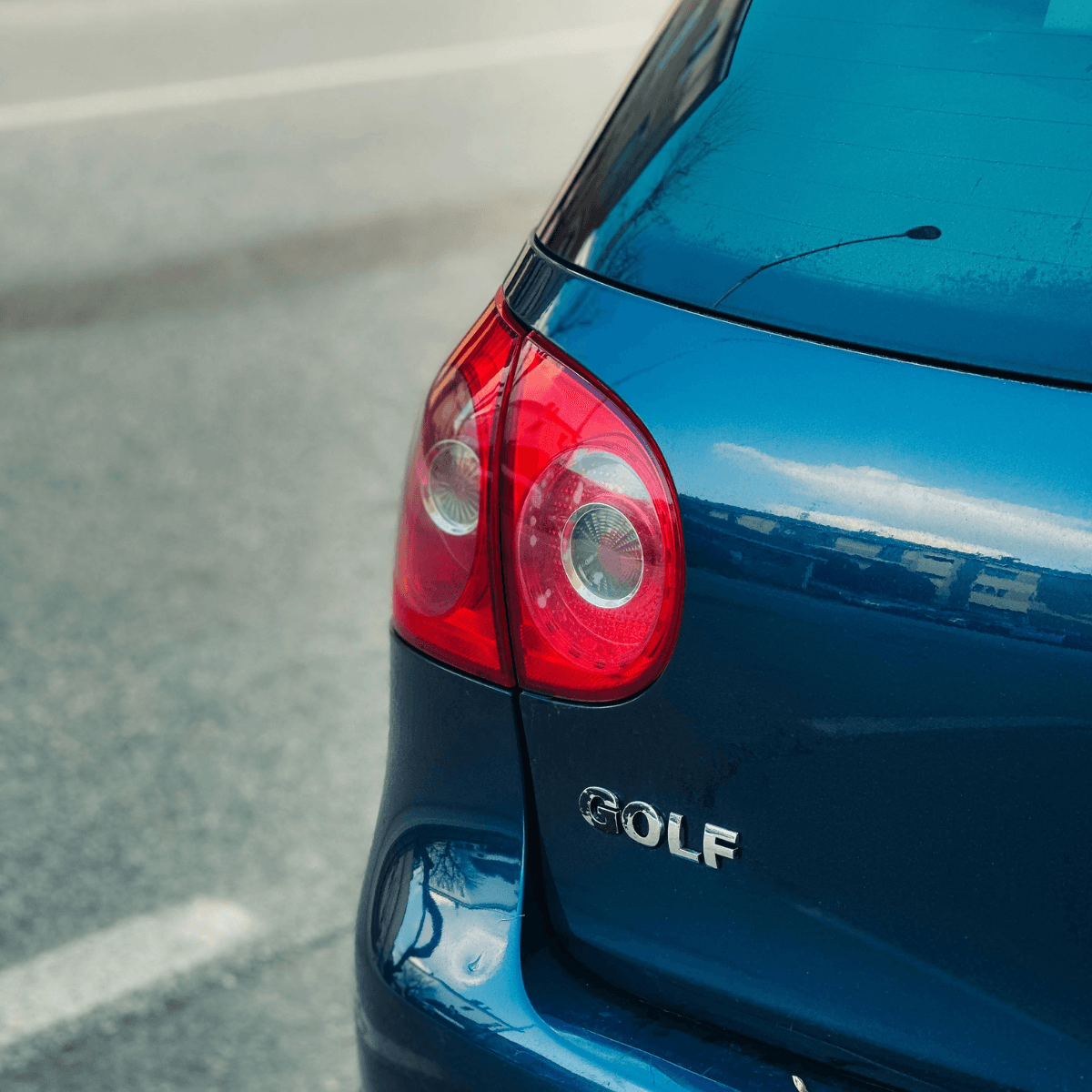 A close-up of the rear corner of a blue Volkswagen Golf, showing the round tail light and chrome Golf badge against a bright outdoor background.