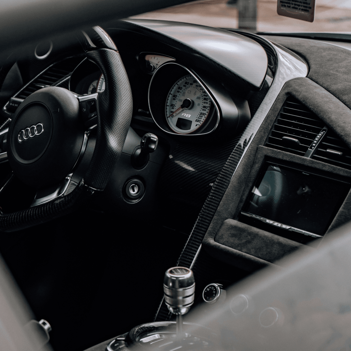 Interior of an Audi R8 showing the carbon fibre trim, Alcantara dashboard, analog speedometer, and manual gear shifter
