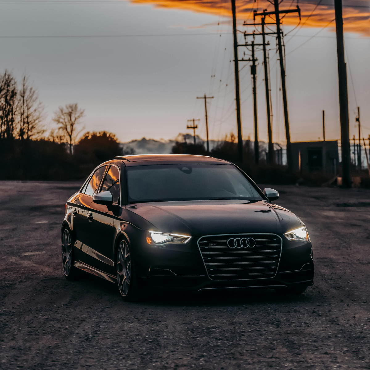 Black Audi S3 sedan with LED headlights on, parked on gravel at dusk with power lines and a dramatic orange and purple sunset sky in the background