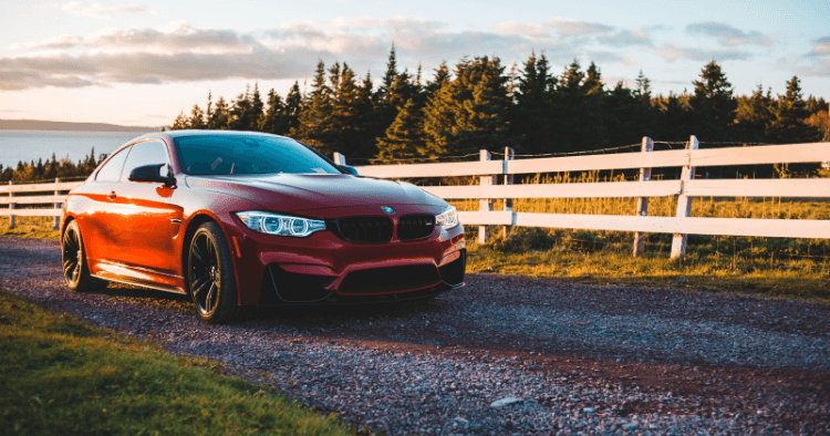 Red BMW coupe with black wheels parked on a gravel road beside a white fence at sunset