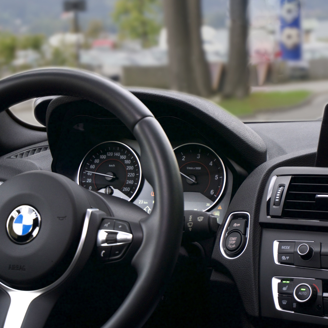 Interior view of a BMW showing the steering wheel, instrument cluster with speedometer reading in km/h, tachometer, start/stop engine button, and climate control panel, with a tree-lined street visible through the windshield.