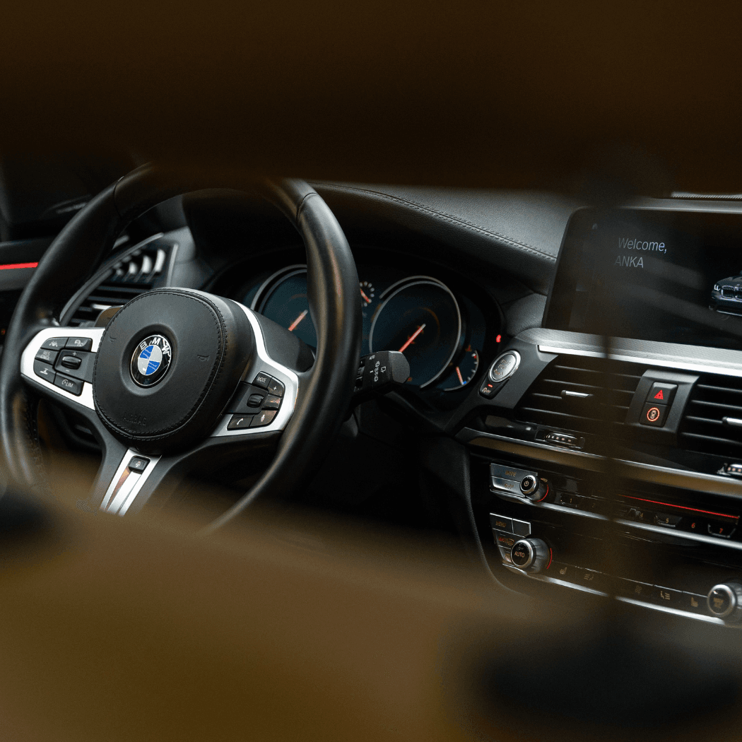 BMW interior shot from the passenger side showing the M Sport steering wheel, glowing instrument gauges with red accents, centre console controls, and an infotainment screen displaying "Welcome, ANKA."
