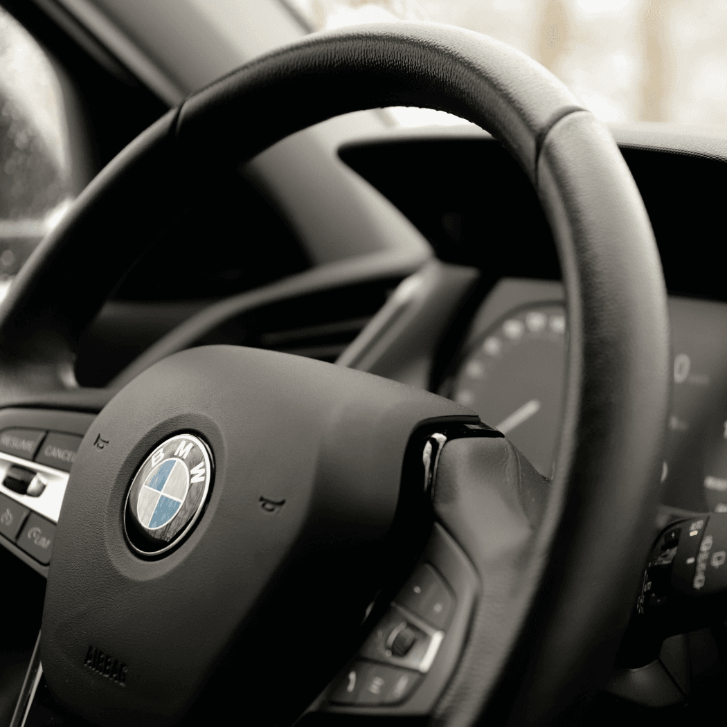  Close-up of a BMW steering wheel in black leather showing the BMW roundel, cruise control buttons, and paddle shifter, with the instrument cluster softly visible in the background.