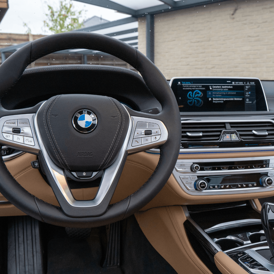 BMW 7 Series interior showing the full steering wheel with brushed silver trim, tan leather dashboard, and a large iDrive infotainment screen displaying Dutch-language vehicle notifications.
