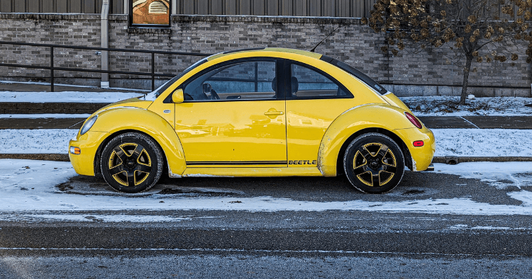Yellow Volkswagen New Beetle with gold wheels parked on a snowy street beside a brick building