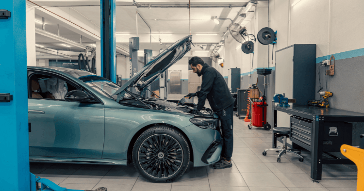 Mechanic inspecting the engine bay of a light blue Mercedes sedan on a lift.