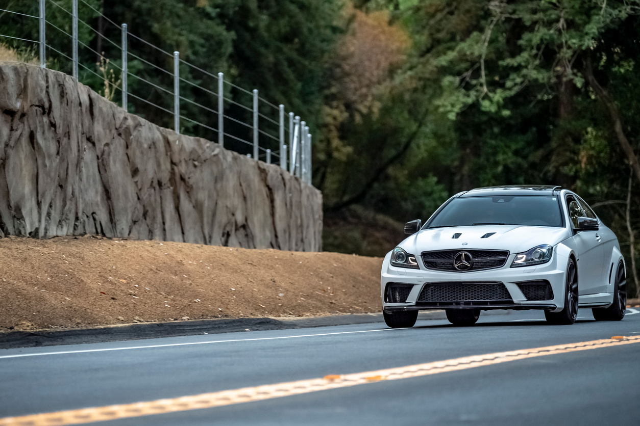 White Mercedes-Benz sports car driving on a road beside a rock wall and surrounded by trees