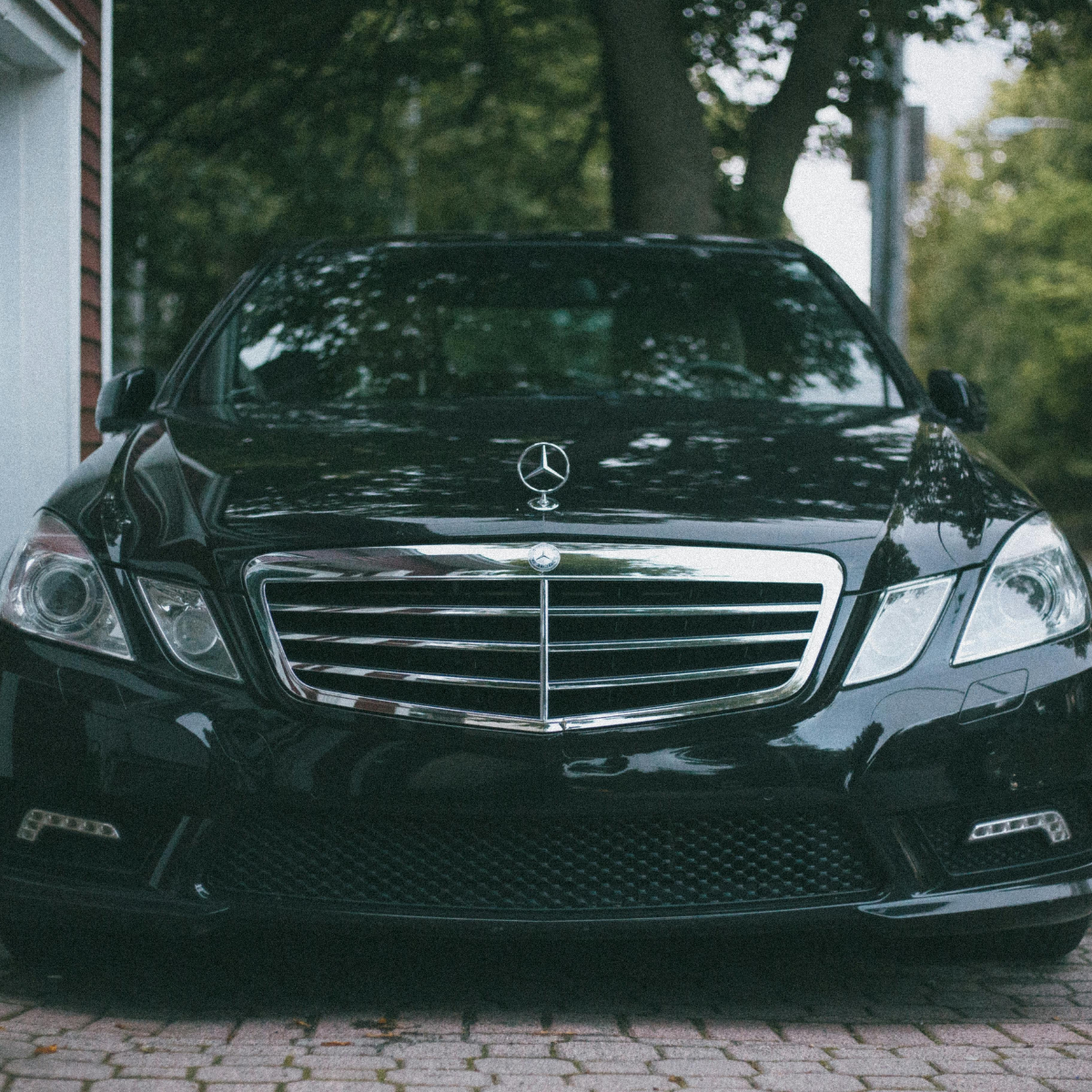 A black Mercedes-Benz E-Class sedan parked on a cobblestone driveway, shot head-on with the chrome grille and hood ornament prominent, surrounded by trees and a brick building.