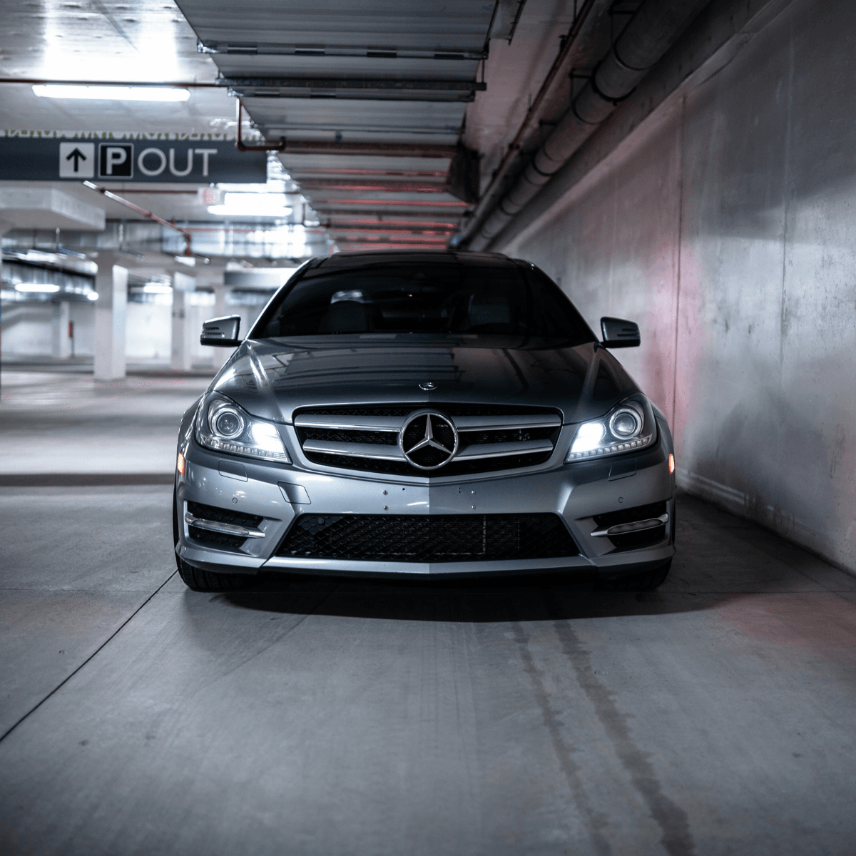 A silver Mercedes-Benz C-Class coupe with its headlights on, photographed head-on inside a parkade with a parking exit sign visible overhead.
