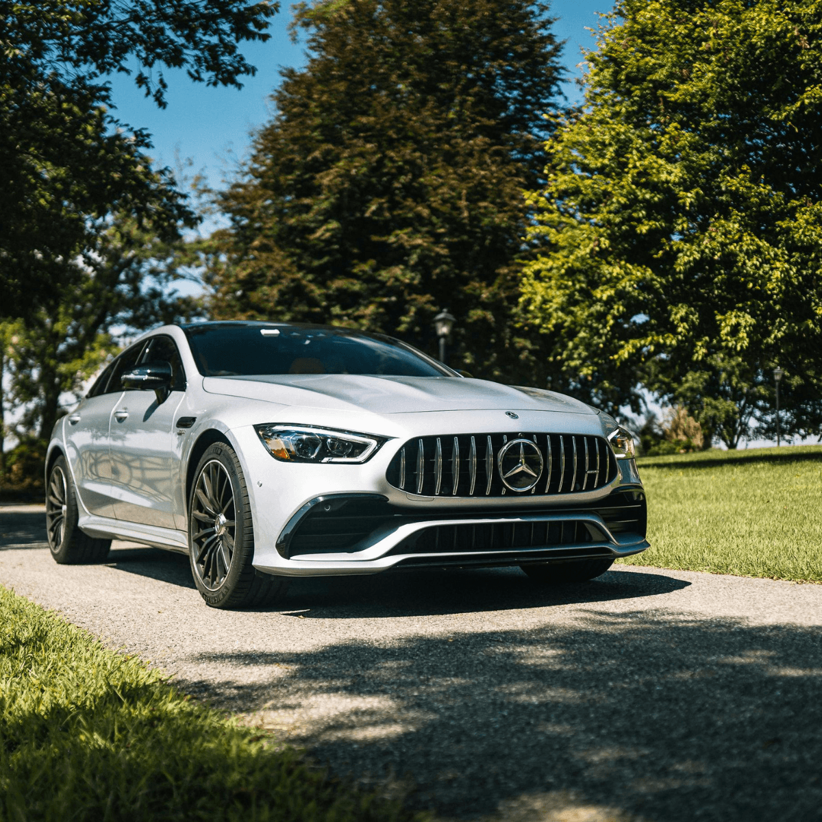 A silver Mercedes-Benz AMG GT 4-Door Coupe parked on a driveway, shot from a low front-quarter angle with bright blue sky and mature trees in the background.