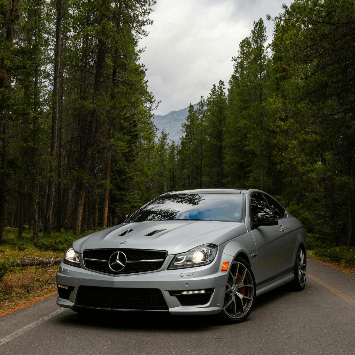 A silver Mercedes-Benz C63 AMG coupe parked on a forest road, shot head-on with tall pine trees and a mountain visible in the background under an overcast sky.