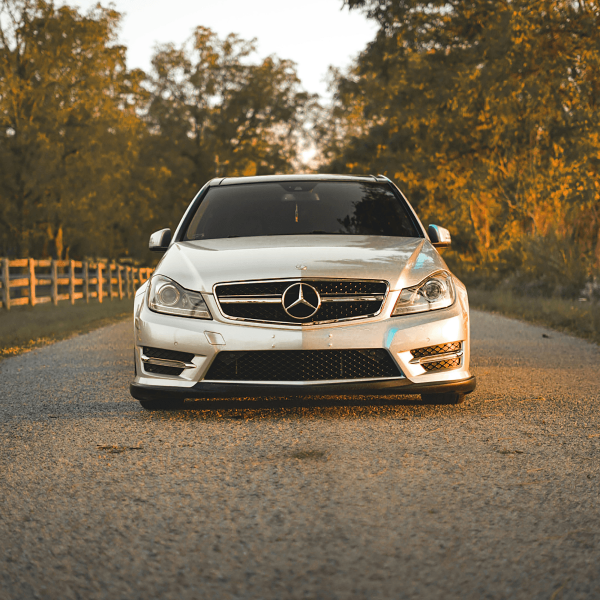 A white Mercedes-Benz C-Class sedan facing the camera head-on on a gravel road, bathed in warm golden-hour light with a wooden fence and autumn trees in the background.