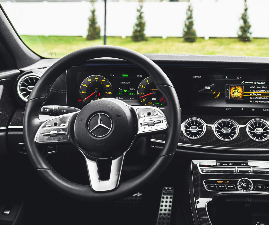 Interior view of a Mercedes-Benz steering wheel with integrated controls, flanked by a dual digital instrument cluster and infotainment screen displaying a SiriusXM radio menu.