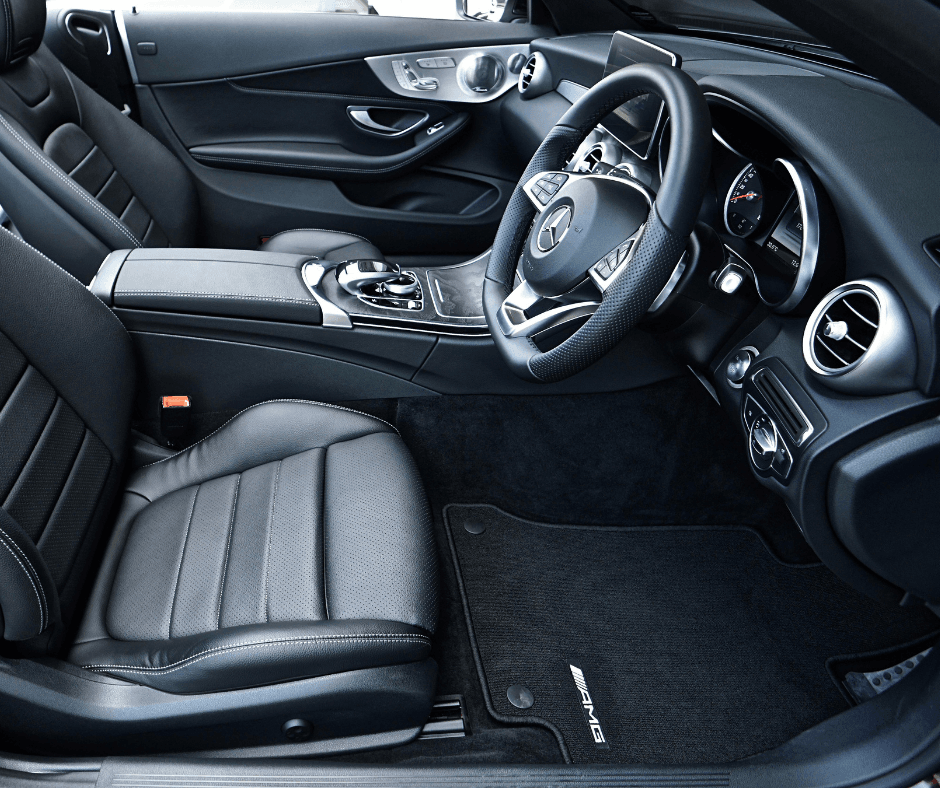 Wide interior view of a Mercedes-AMG C-Class showing black perforated leather seats with white contrast stitching, an AMG floor mat, centre console, and steering wheel.