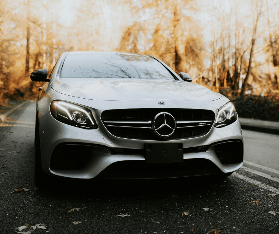 Front view of a silver Mercedes-AMG E-Class sedan parked on a road surrounded by autumn trees, with the three-pointed star badge prominently centred on the front grille.