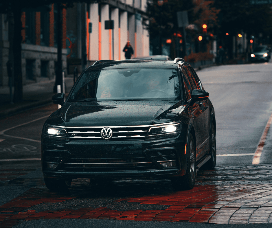 A black Volkswagen Tiguan SUV driving on a wet city street at dusk, with illuminated headlights and a urban backdrop of buildings and traffic lights.