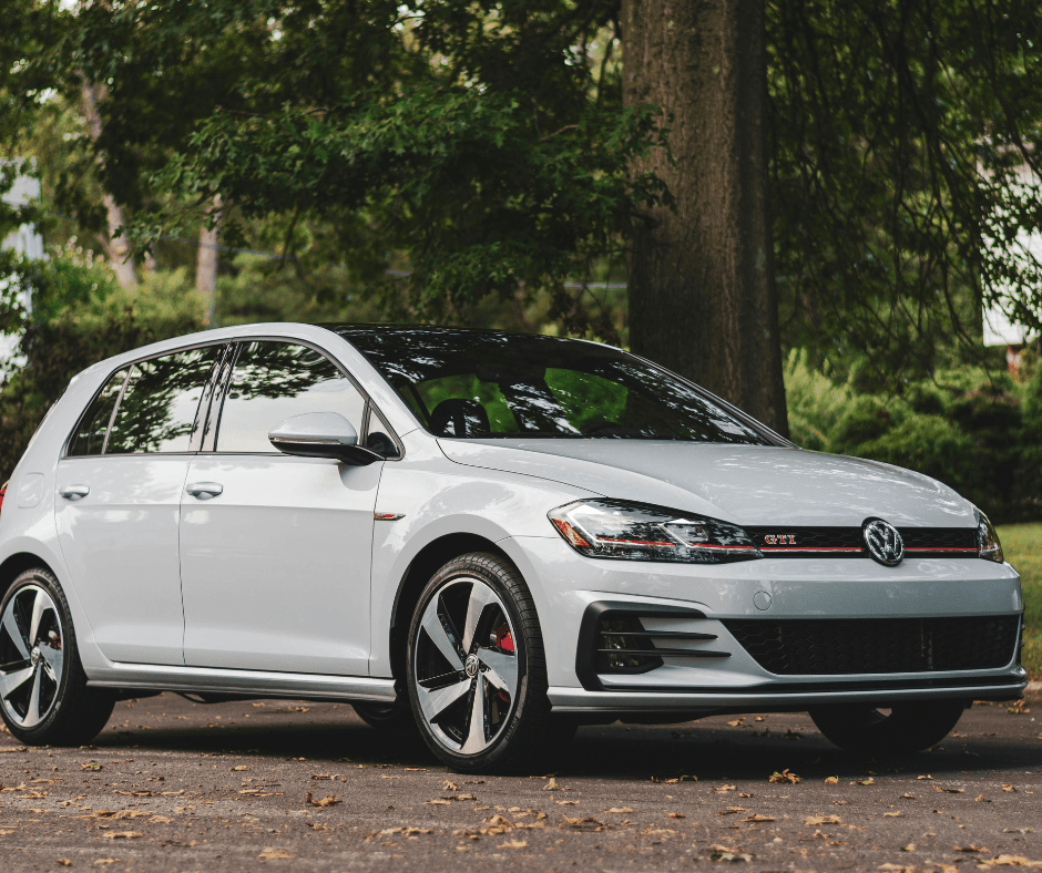 A white Volkswagen Golf GTI parked outdoors on a tree-lined driveway, showing the GTI badge and red accent stripe on the front grille.