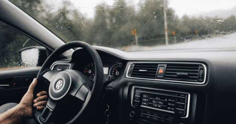 Driver's hand on a Volkswagen steering wheel, rain streaking the windshield ahead.
