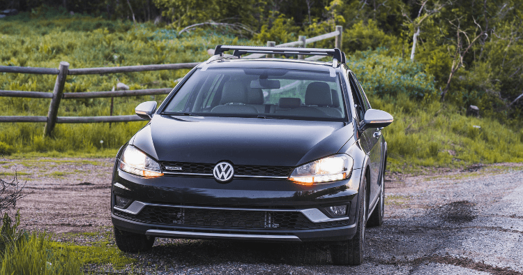 Black Volkswagen Golf Alltrack driving down a gravel road past a wooden fence.