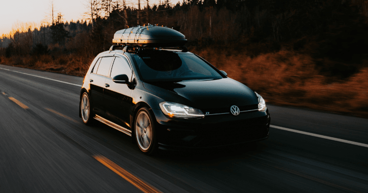 Black Volkswagen Golf with rooftop cargo box driving on a highway at dusk.