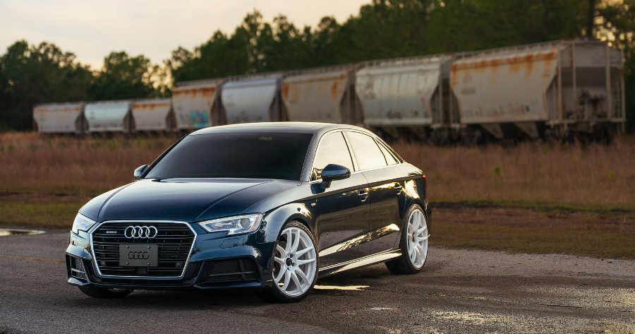 Dark blue Audi A3 sedan with white aftermarket wheels parked near a freight train at sunset