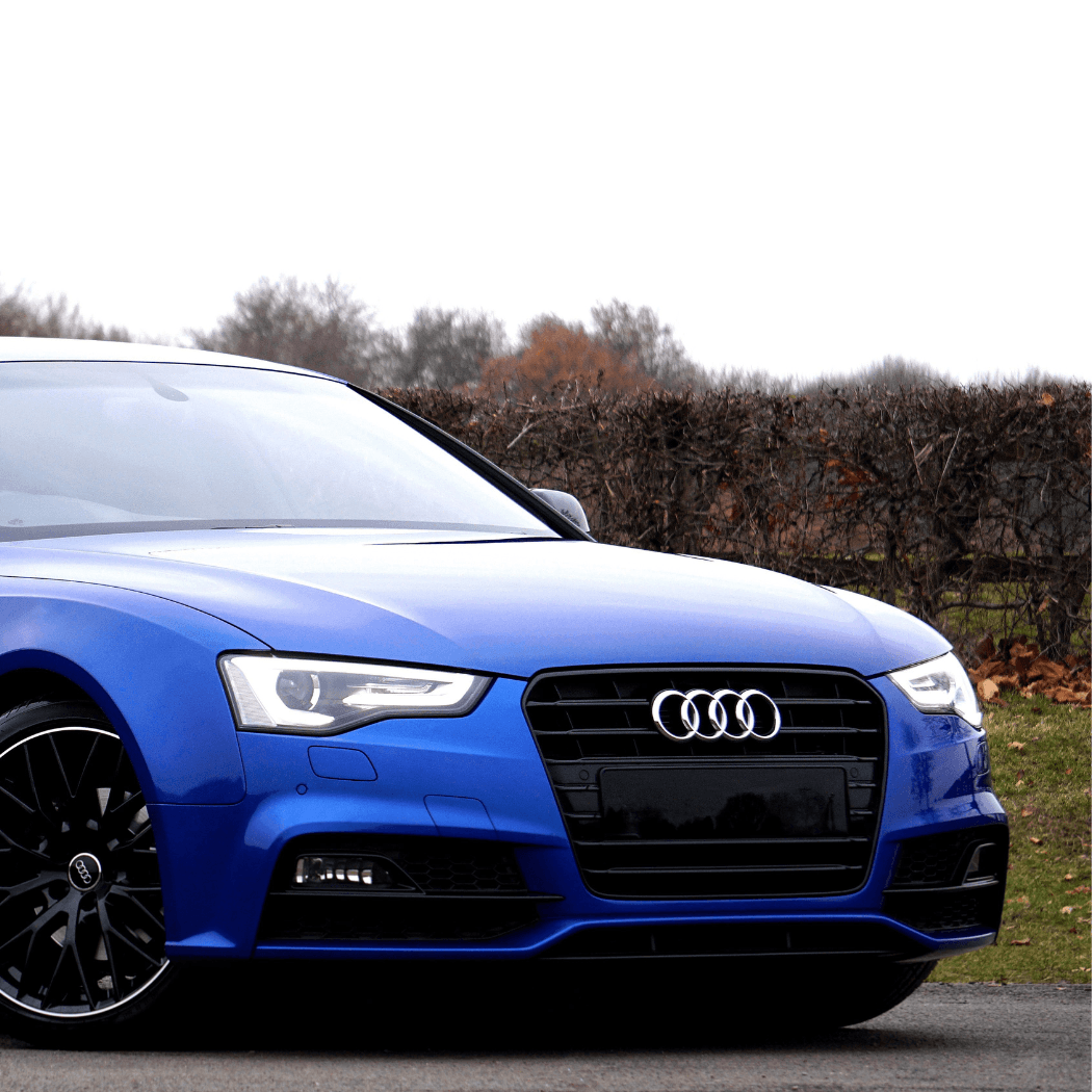 Front corner view of a vivid blue Audi A5 with black alloy wheels, shot against a hedgerow with autumn foliage, highlighting the sculpted hood and LED running lights.