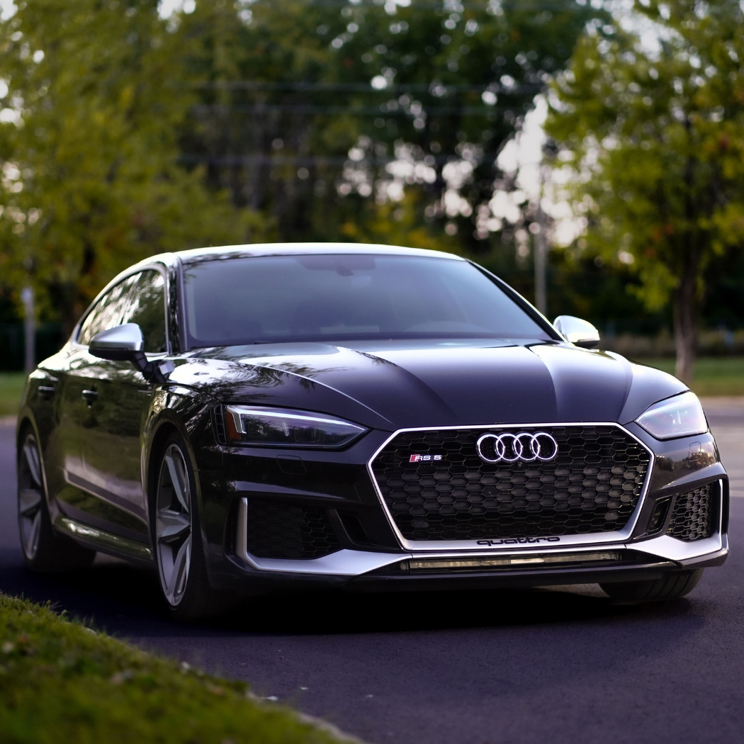 Front exterior of a dark grey Audi RS5 Sportback with quattro all-wheel drive, shot in a leafy suburban setting with the hexagonal grille and RS5 badge clearly visible.