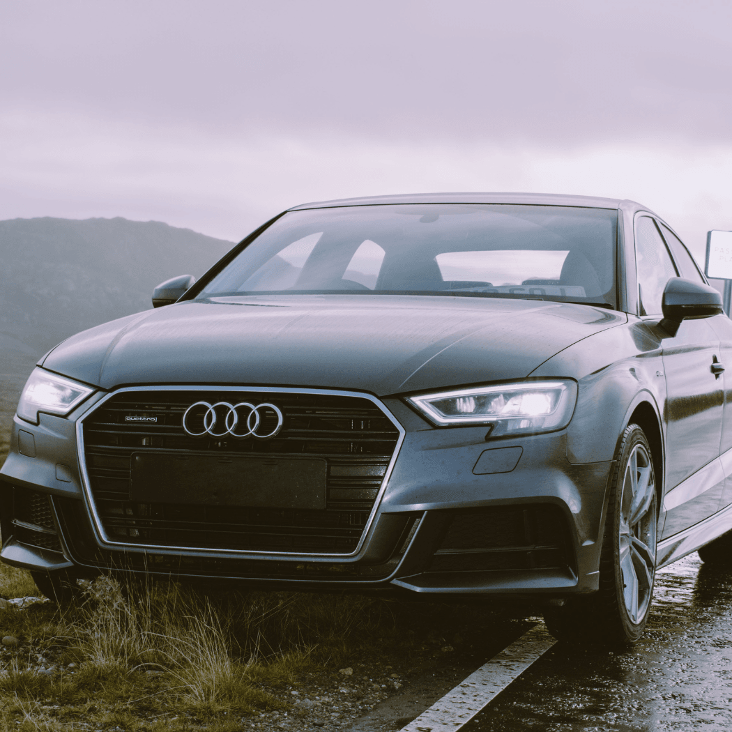 Front exterior of a dark grey Audi A3 quattro parked on a wet mountain road under an overcast sky, with headlights on and rugged highland terrain in the background.