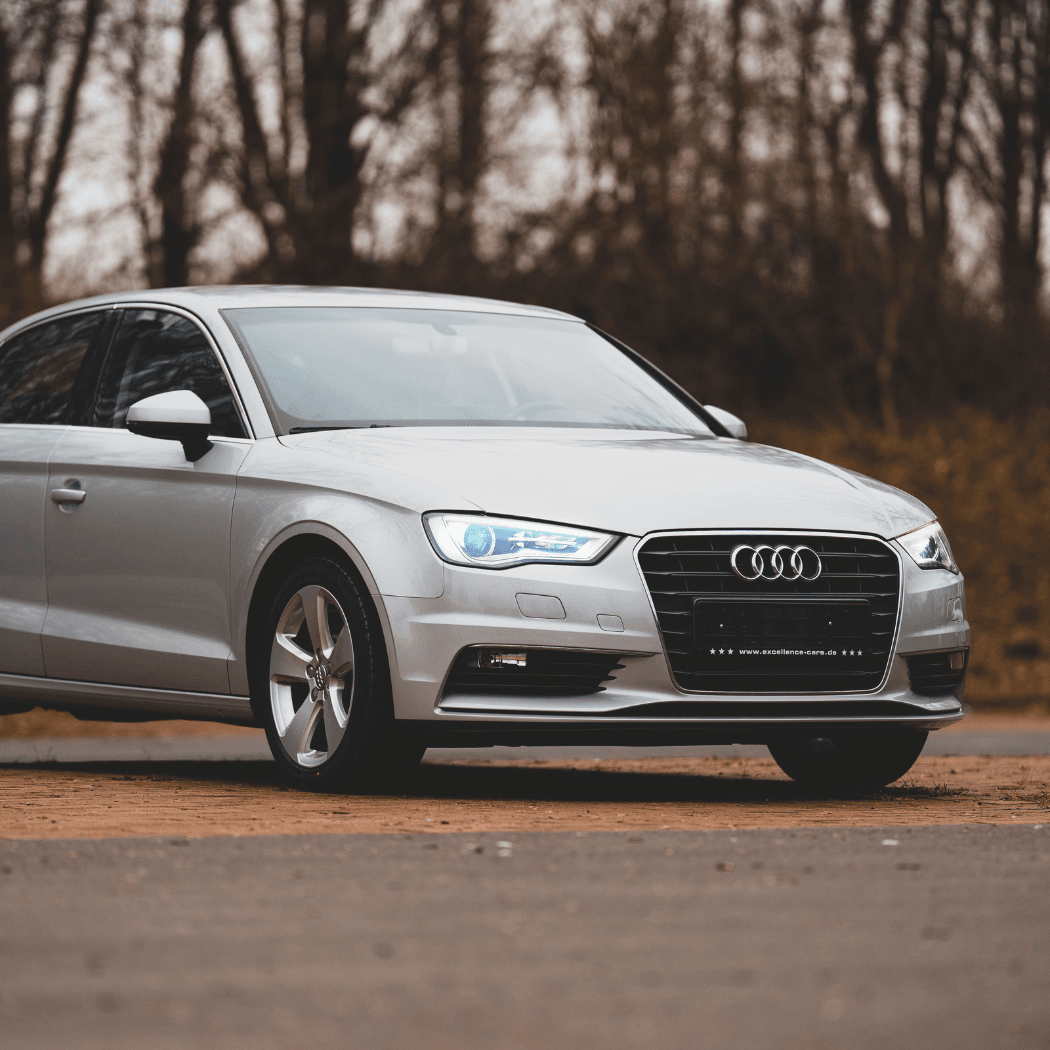 Front three-quarter exterior of a silver Audi A3 sedan with LED daytime running lights, parked on a dirt surface against a backdrop of bare winter trees.