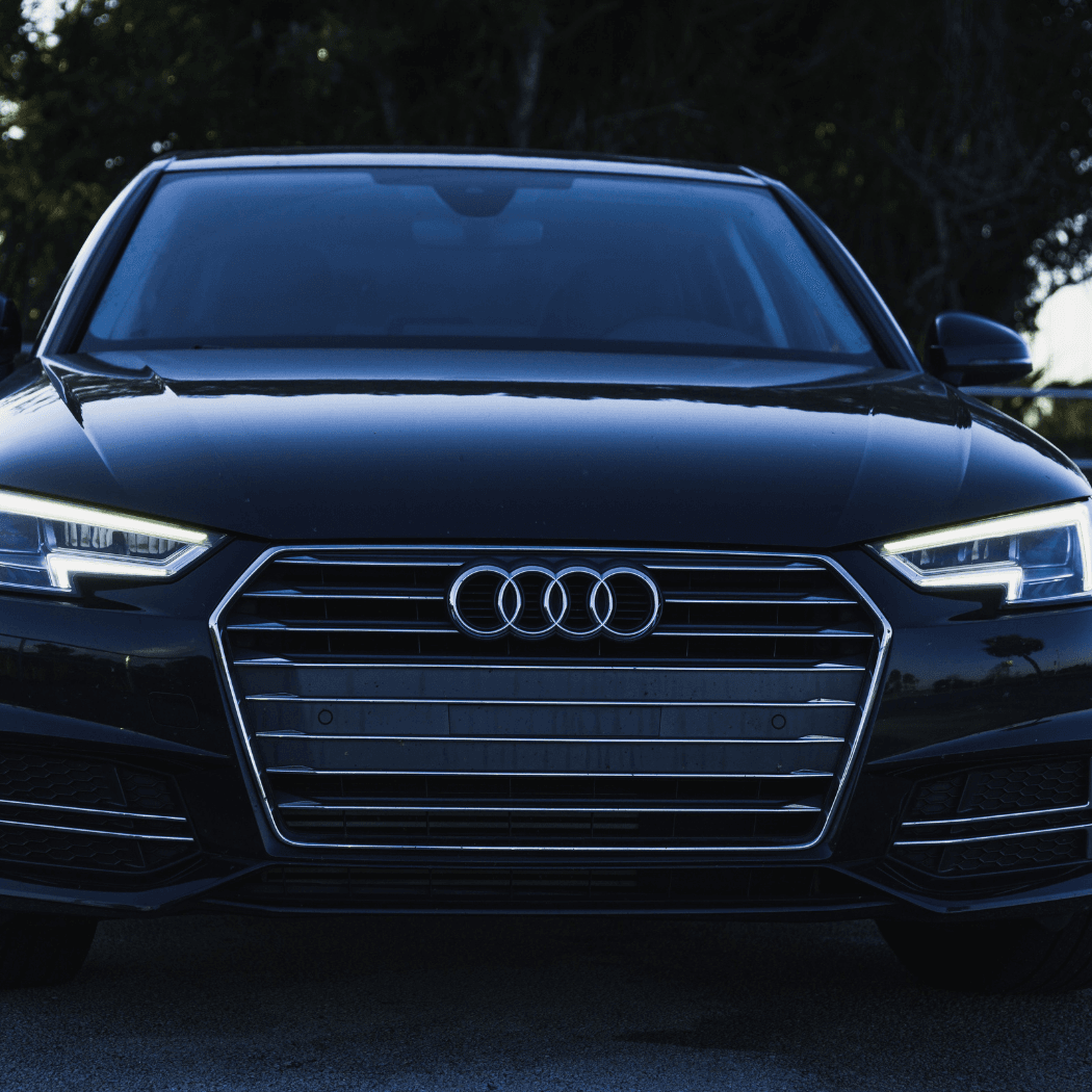 Straight-on front exterior of a black Audi A4 at dusk, with sharp LED headlights illuminated and the chrome four-ring badge centred on the single-frame grille.
